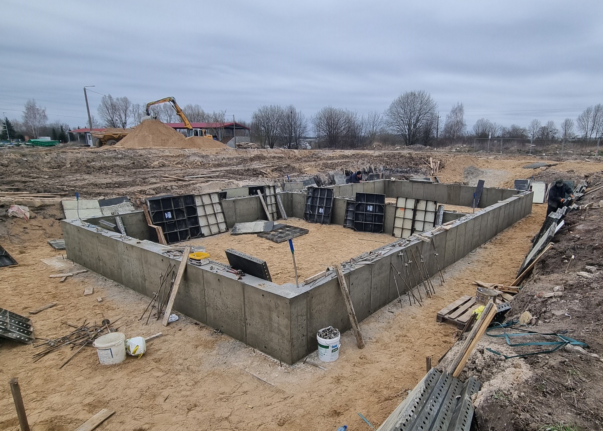 Wylany fundament pod budynek z częściowo zdjętym szalunkiem, widoczne zbrojenia i narzędzia budowlane, w tle koparka i hałda piasku.