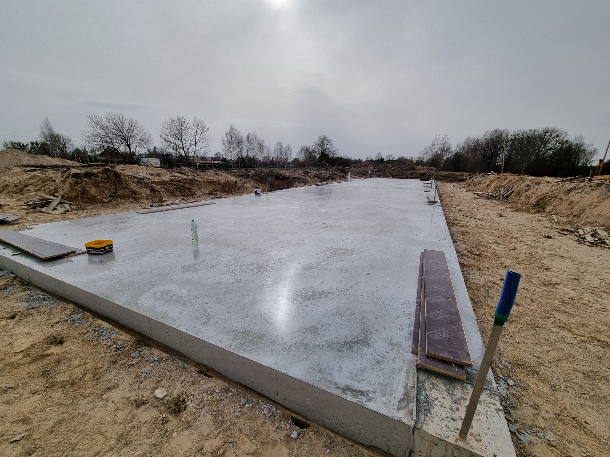 Świeżo wylany, gładki fundament płyty budynku w Orzyszu, z widocznymi deskami szalunkowymi i narzędziem do rozprowadzania betonu, na tle pochmurnego nieba.