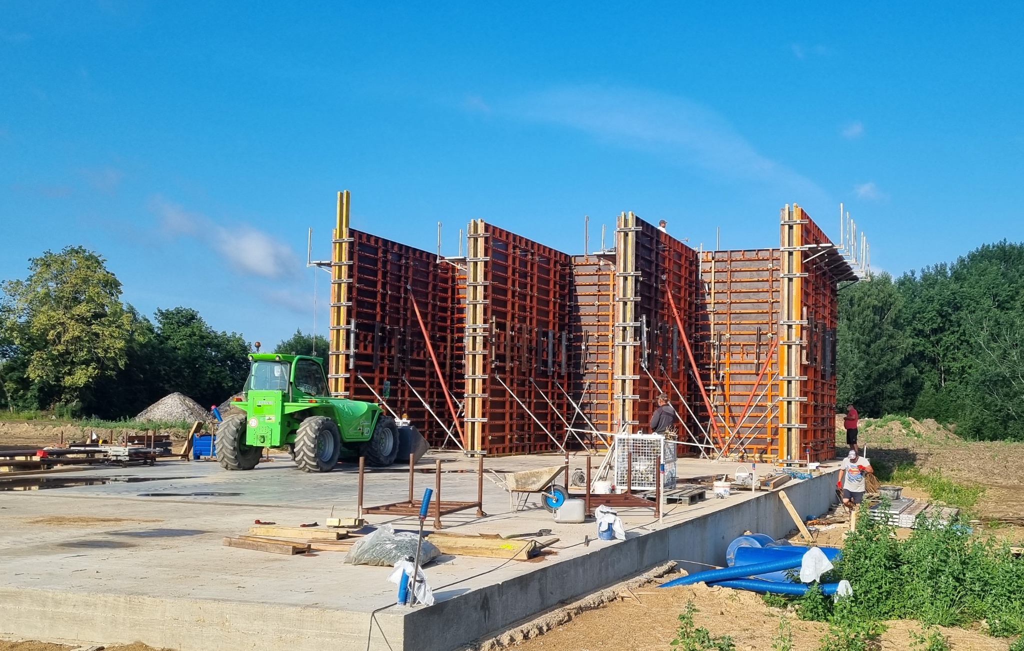 Konstrukcja szalunkowa pod budynek żelbetowy z widocznym zielonym podnośnikiem teleskopowym oraz pracownikami na placu budowy w słoneczny dzień.