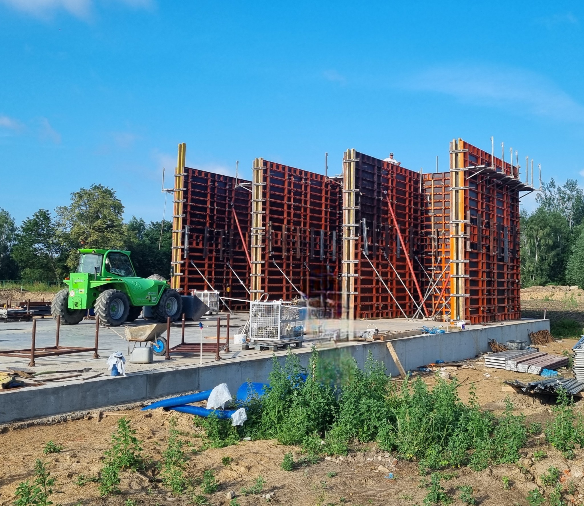 Szare fundamenty z deskowaniem szalunkowym, zielony wózek widłowy Merlo na placu budowy, błękitne niebo.