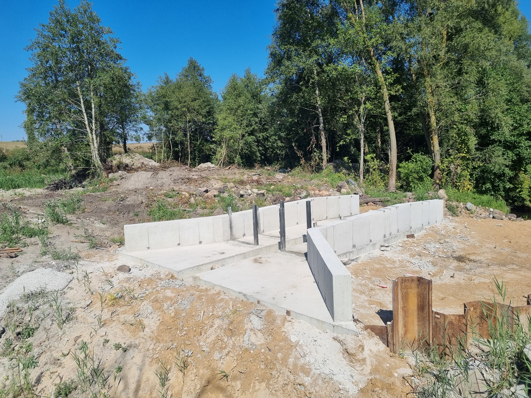 Wylany fundament pod budynek z widocznymi stalowymi elementami szalunku w gruncie, otoczony drzewami i ziemią.