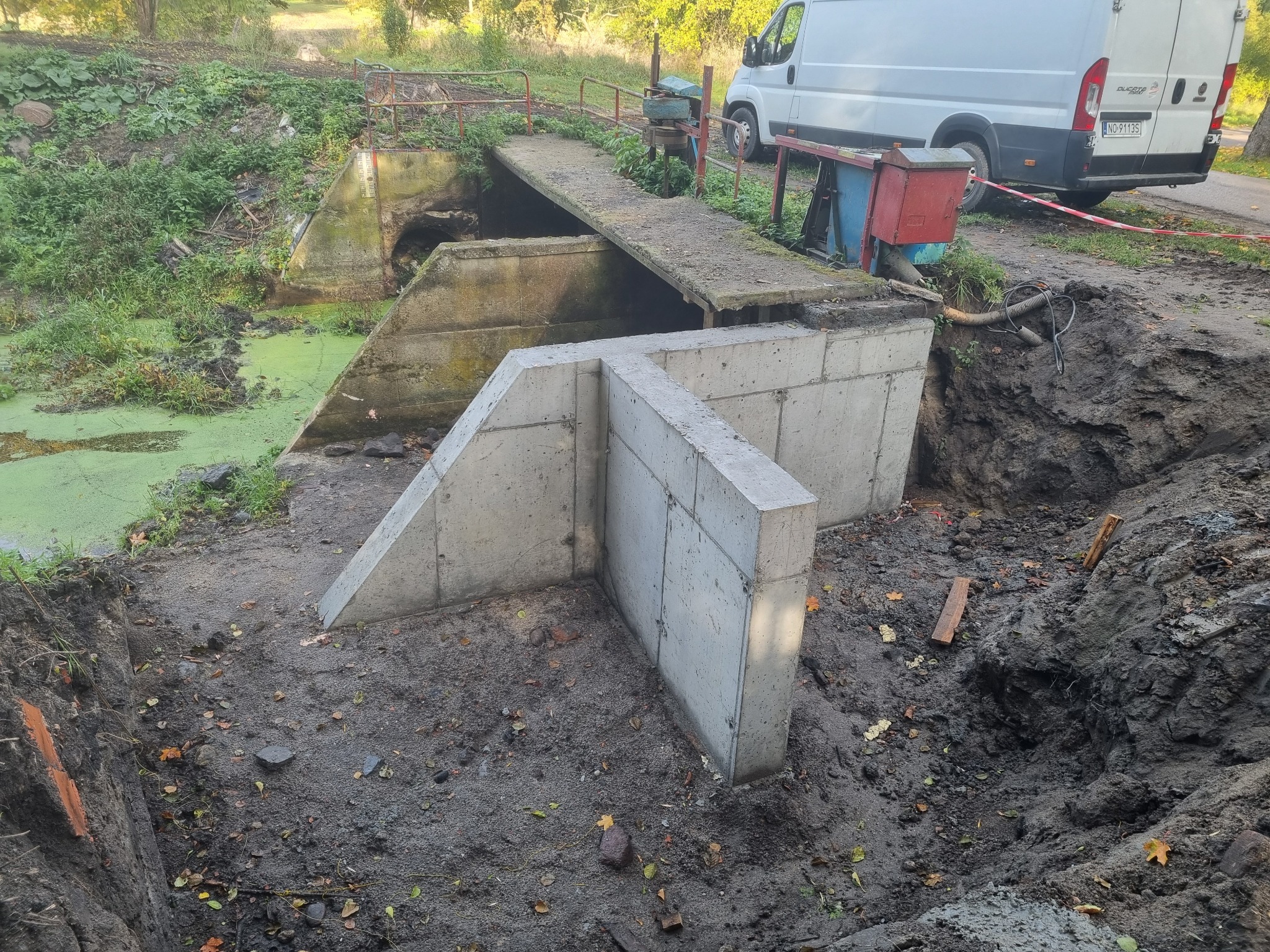 Nowy żelbetowy element konstrukcyjny w korycie rzeki, obok starego mostku z zarośniętą roślinnością i samochodem dostawczym w tle.