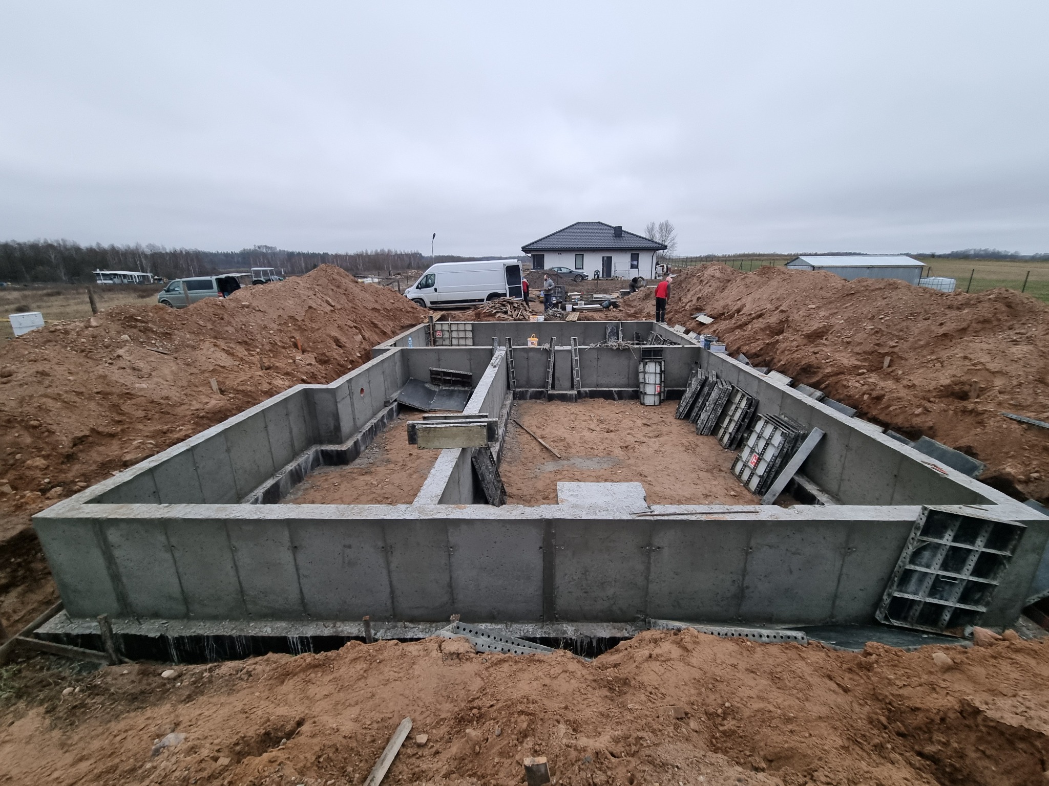 Zalany betonem fundament domu jednorodzinnego, widoczne szalunki i wykop ziemny, w tle biały bus i budynek mieszkalny.