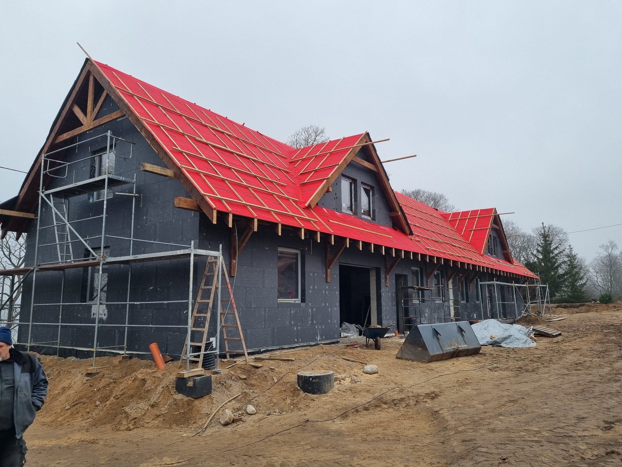 Budowa domu: czarna elewacja z izolacją, czerwona membrana dachowa na drewnianej konstrukcji, rusztowania, teren budowy w trakcie prac.