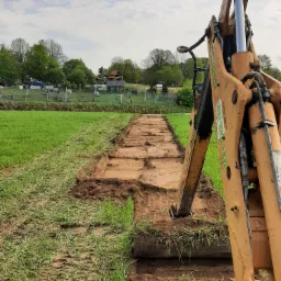 Żółta koparka wykopuje wąski rów w zielonym polu, widoczne świeżo wykopane fragmenty ziemi i fragmenty trawy na brzegach, w tle zabudowania wiejskie pod błękitnym niebem.