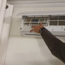 Dariusz Andreasik - Biały klimatyzator ścienny z otwartą klapą, widoczne filtry, ręka mężczyzny w brązowej bluzie z zegarkiem reguluje położenie kratki filtra.