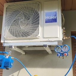 Dariusz Andreasik - Zewnętrzna jednostka klimatyzacji Haier zamontowana na ścianie budynku, podłączona do pompy próżniowej i manometrów za pomocą niebieskich przewodów podczas serwisu.