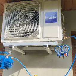 Zewnętrzna jednostka klimatyzacji Haier zamontowana na ścianie budynku, podłączona do pompy pr&oacute;żniowej i manometr&oacute;w za pomocą niebieskich przewod&oacute;w podczas serwisu.