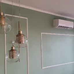 Biała jednostka klimatyzacyjna zamontowana na ścianie w pokoju z pastelowo zielonymi ścianami, ozdobionymi białymi listwami, widoczne trzy wiszące lampy z ażurowymi kloszami i okno.