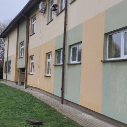 Dariusz Andreasik - Zewnętrzne jednostki klimatyzacyjne zamontowane na elewacji budynku pomalowanej w pasy w odcieniach beżu i zieleni. Widoczne okna z białymi ramami, rynny i trawnik.