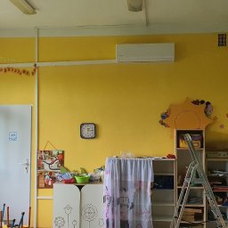 Dariusz Andreasik - Wnętrze sali przedszkolnej z żółtymi ścianami, klimatyzatorem na ścianie, dekoracjami i meblami dziecięcymi, drabina rozstawiona w pobliżu regałów.