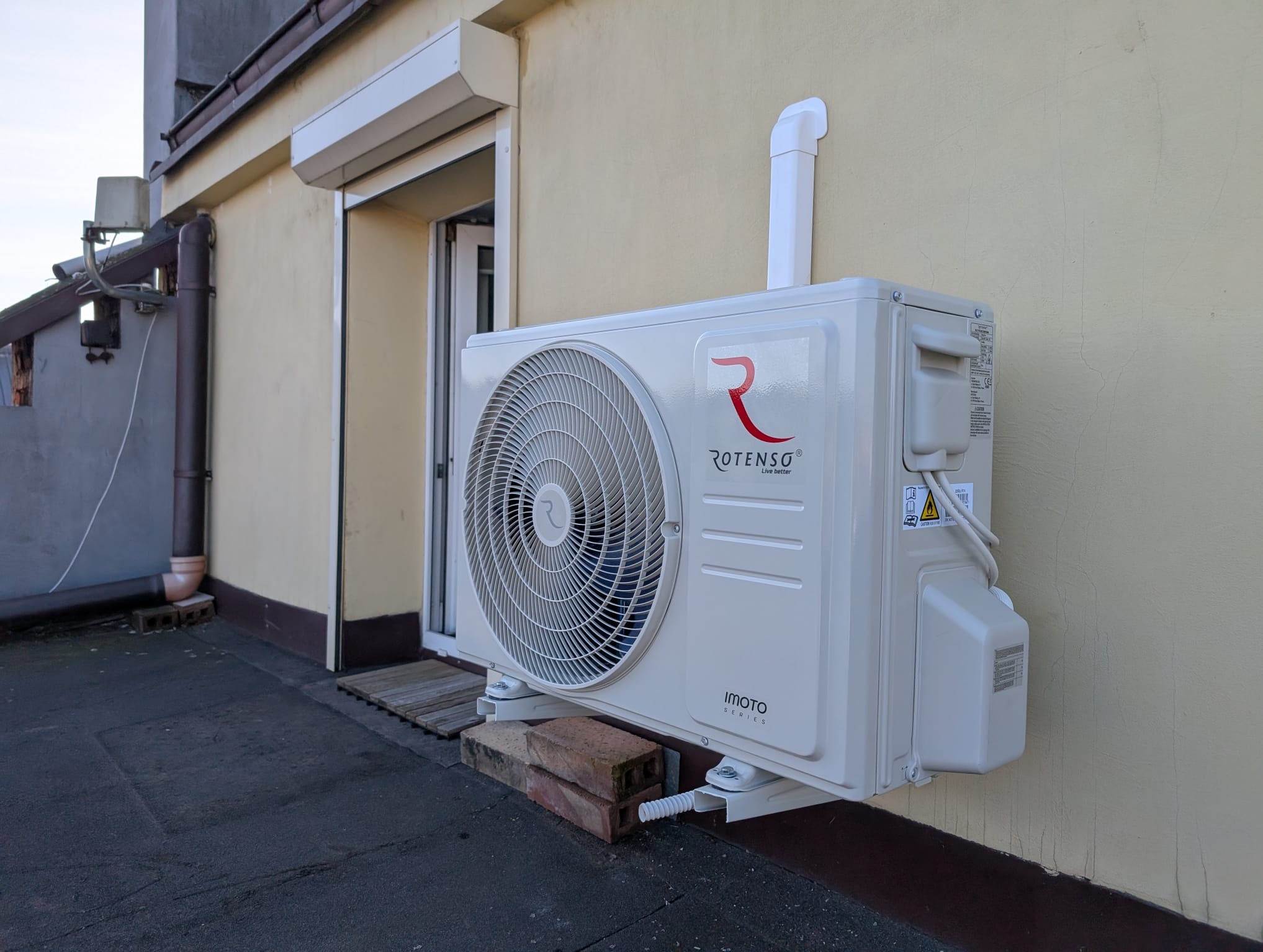 Zewnętrzna jednostka klimatyzacji Rotenso Imoto Series zamontowana na dachu budynku, wsparta na cegłach, z widoczną rurą odprowadzającą skropliny.