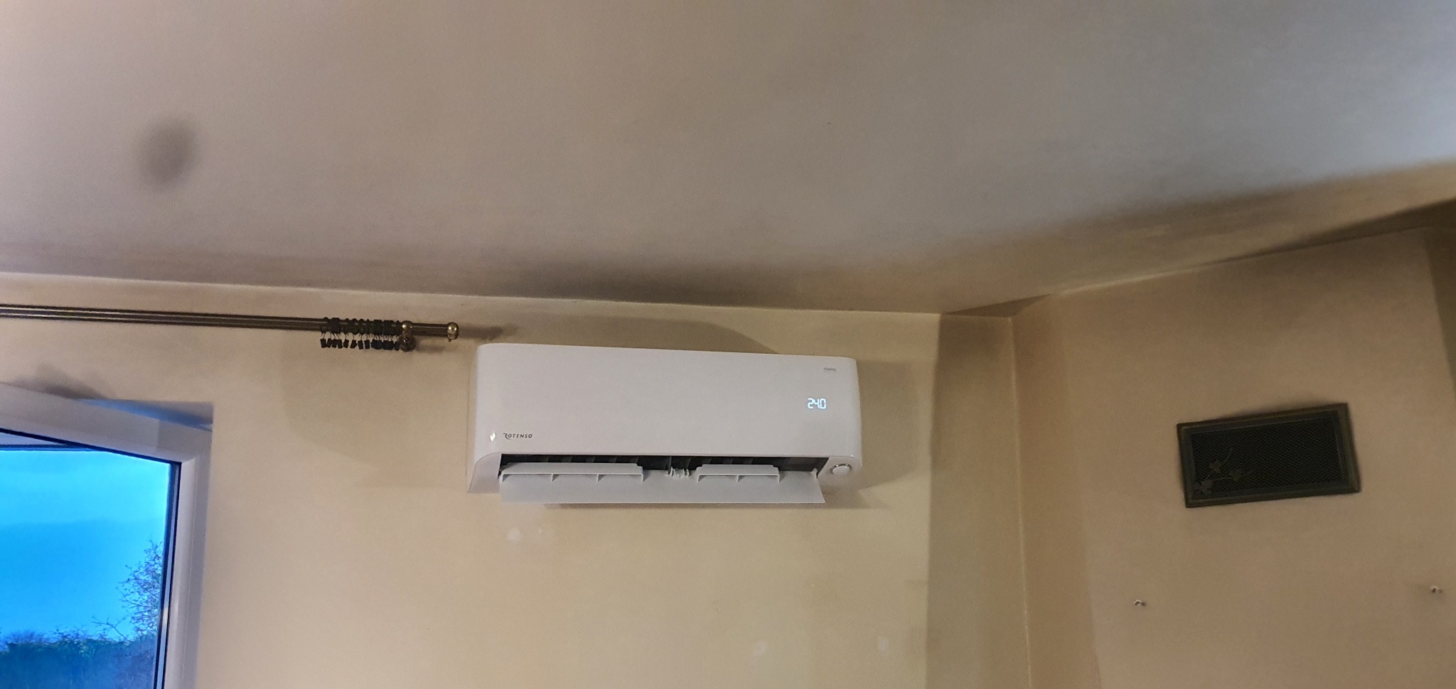 Biały klimatyzator Rotenso zamontowany na ścianie w jasnym pomieszczeniu, widoczny wyświetlacz z temperaturą 24.0 stopnie Celsjusza, obok okno i stara kratka wentylacyjna.