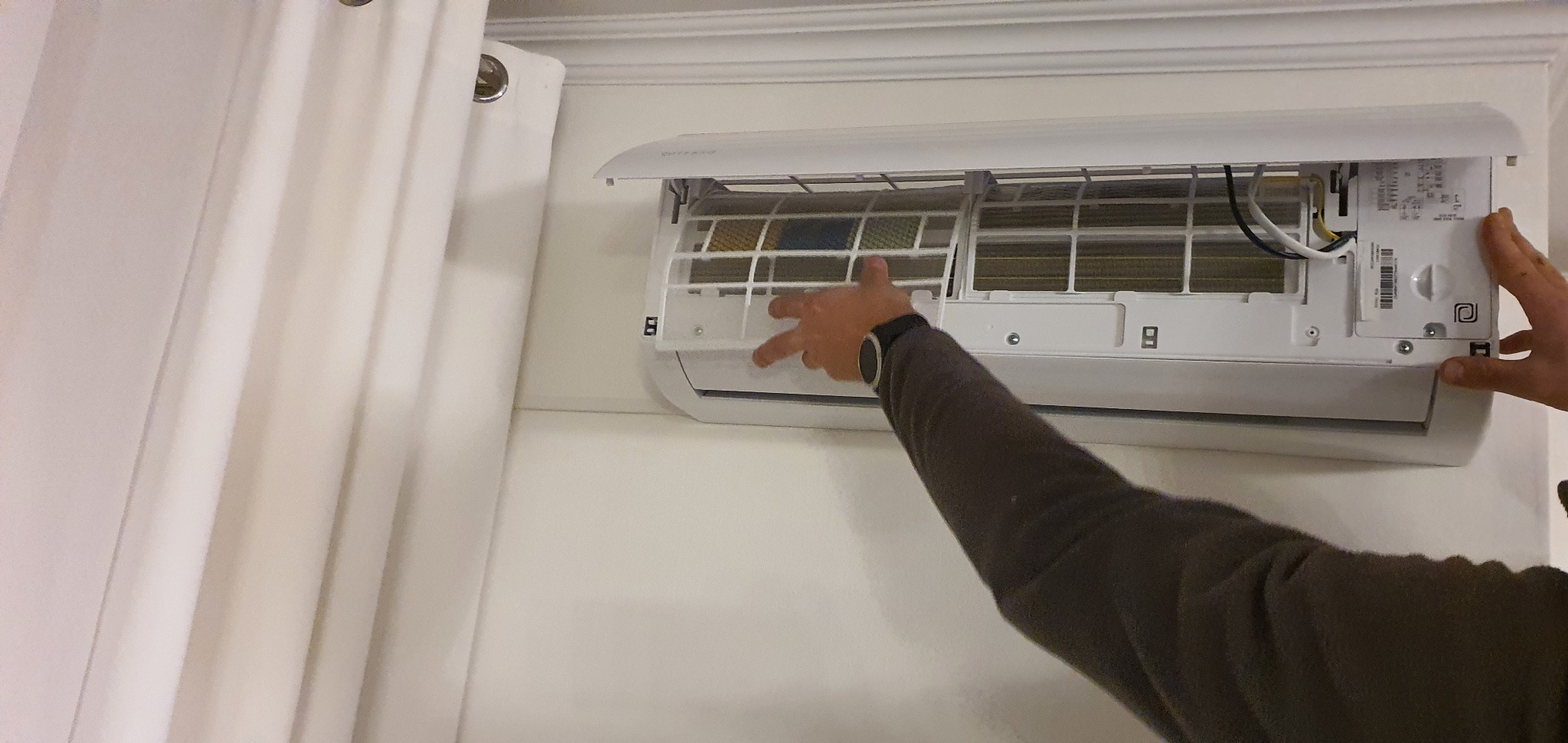 Biały klimatyzator ścienny z otwartą klapą, widoczne filtry, ręka mężczyzny w brązowej bluzie z zegarkiem reguluje położenie kratki filtra.