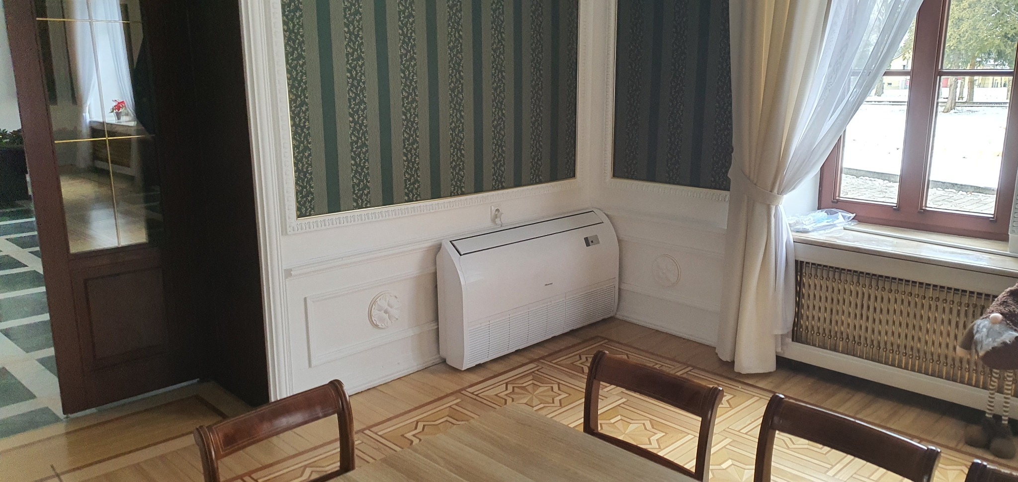 Biała jednostka klimatyzacyjna podłogowa Hisense w pomieszczeniu z dekoracyjnymi listwami ściennymi, wzorzystą tapetą i drewnianą podłogą w geometryczny wzór, widok z perspektywy stołu z krzesłami.