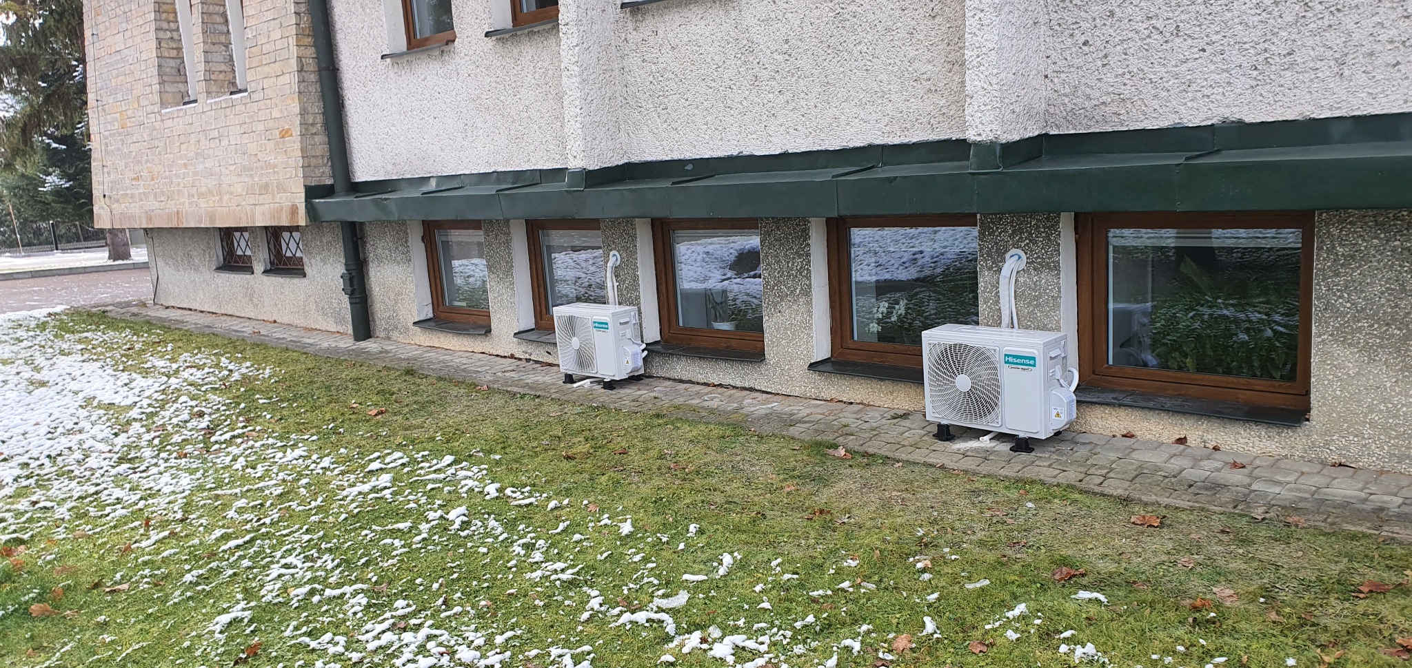 Dwa zewnętrzne agregaty klimatyzacji marki Hisense ustawione na trawniku z resztkami śniegu, pod oknami budynku o szarej elewacji.
