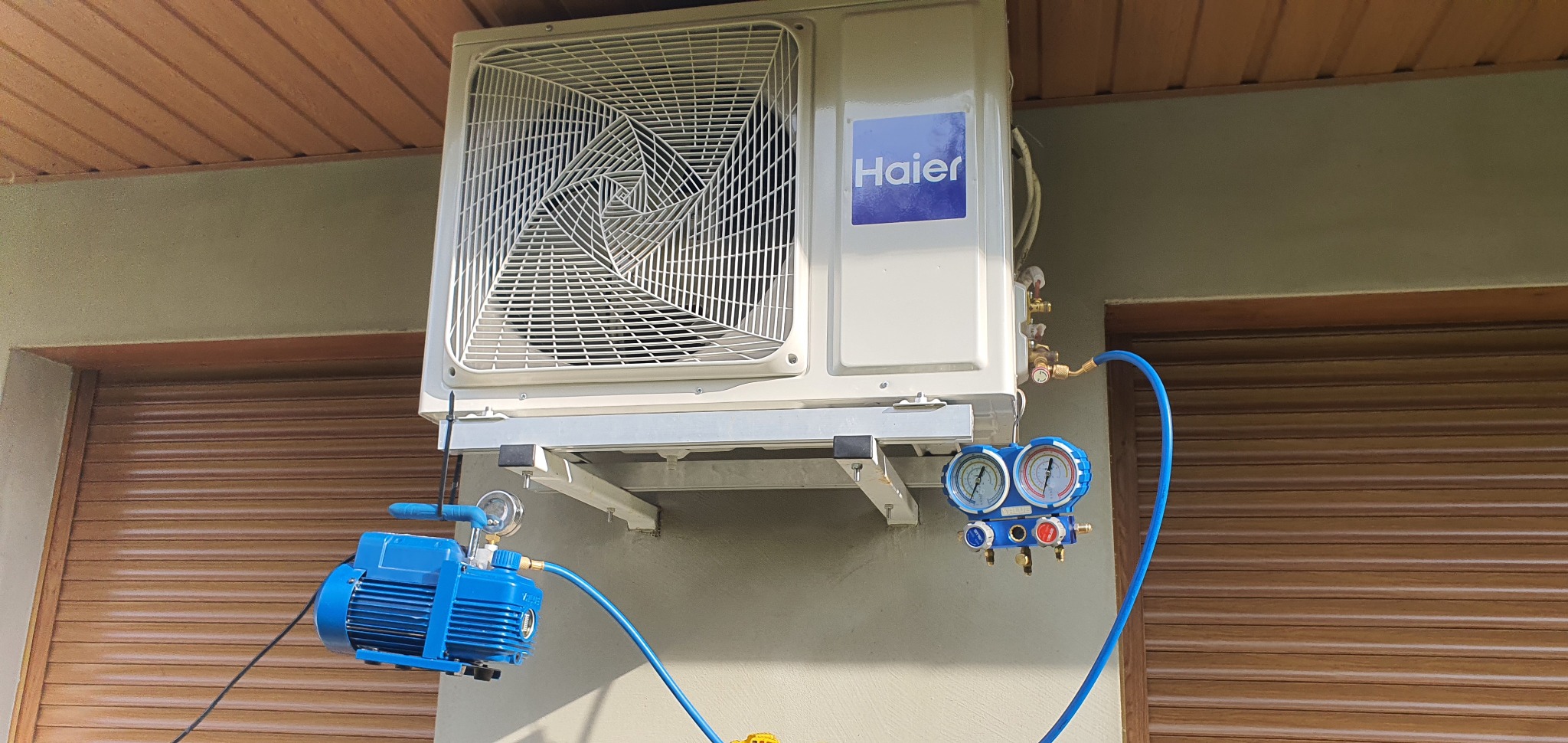 Zewnętrzna jednostka klimatyzacji Haier zamontowana na ścianie budynku, podłączona do pompy próżniowej i manometrów za pomocą niebieskich przewodów podczas serwisu.