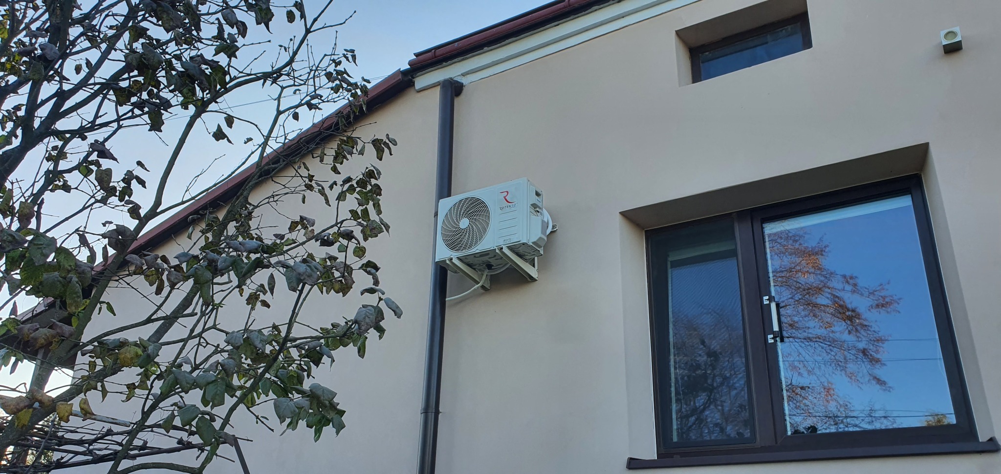 Zewnętrzna jednostka klimatyzacji Rotenso zamontowana na elewacji domu jednorodzinnego, widoczny fragment drzewa i okna z odbijającym się niebem.