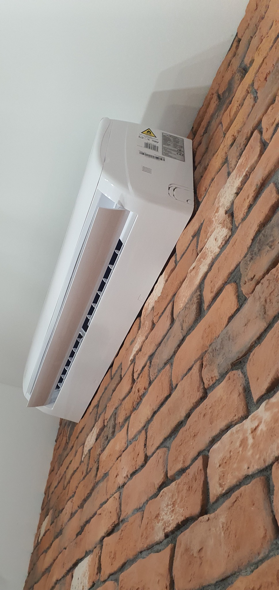 Biały klimatyzator ścienny zamontowany na ścianie z cegły dekoracyjnej, widok z dołu pod kątem.