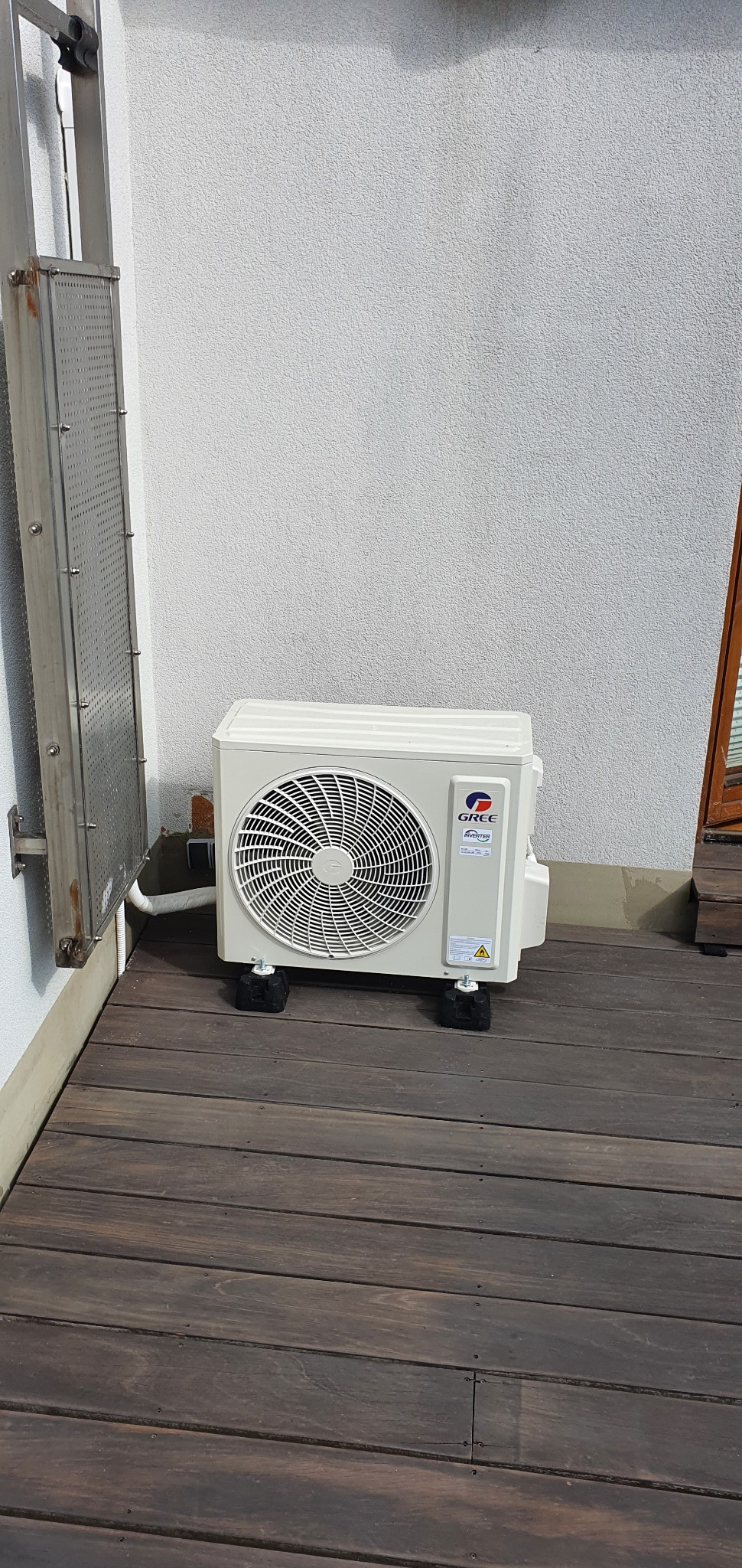 Zewnętrzna jednostka klimatyzacji marki Gree, umieszczona na drewnianym tarasie, z widocznym metalowym koszem na ścianie.