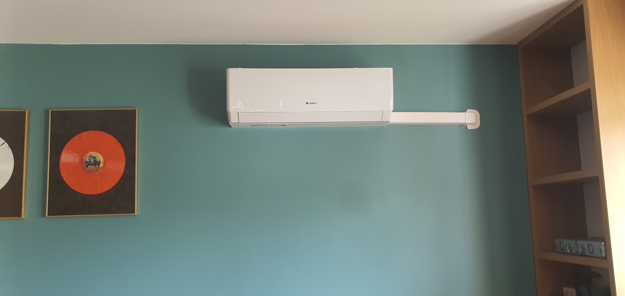 Biała klimatyzacja Gree zamontowana na turkusowej ścianie w pokoju z półkami i obrazami winyli.