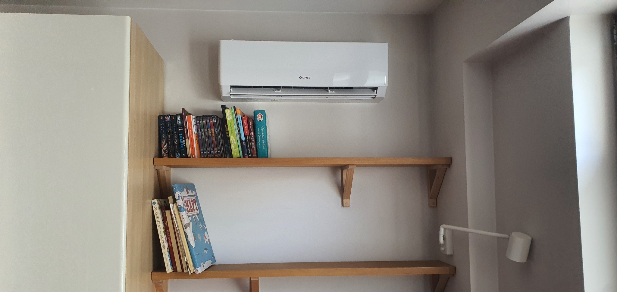 Biały klimatyzator marki Gree zamontowany w pomieszczeniu nad półkami z książkami, w neutralnej kolorystyce.