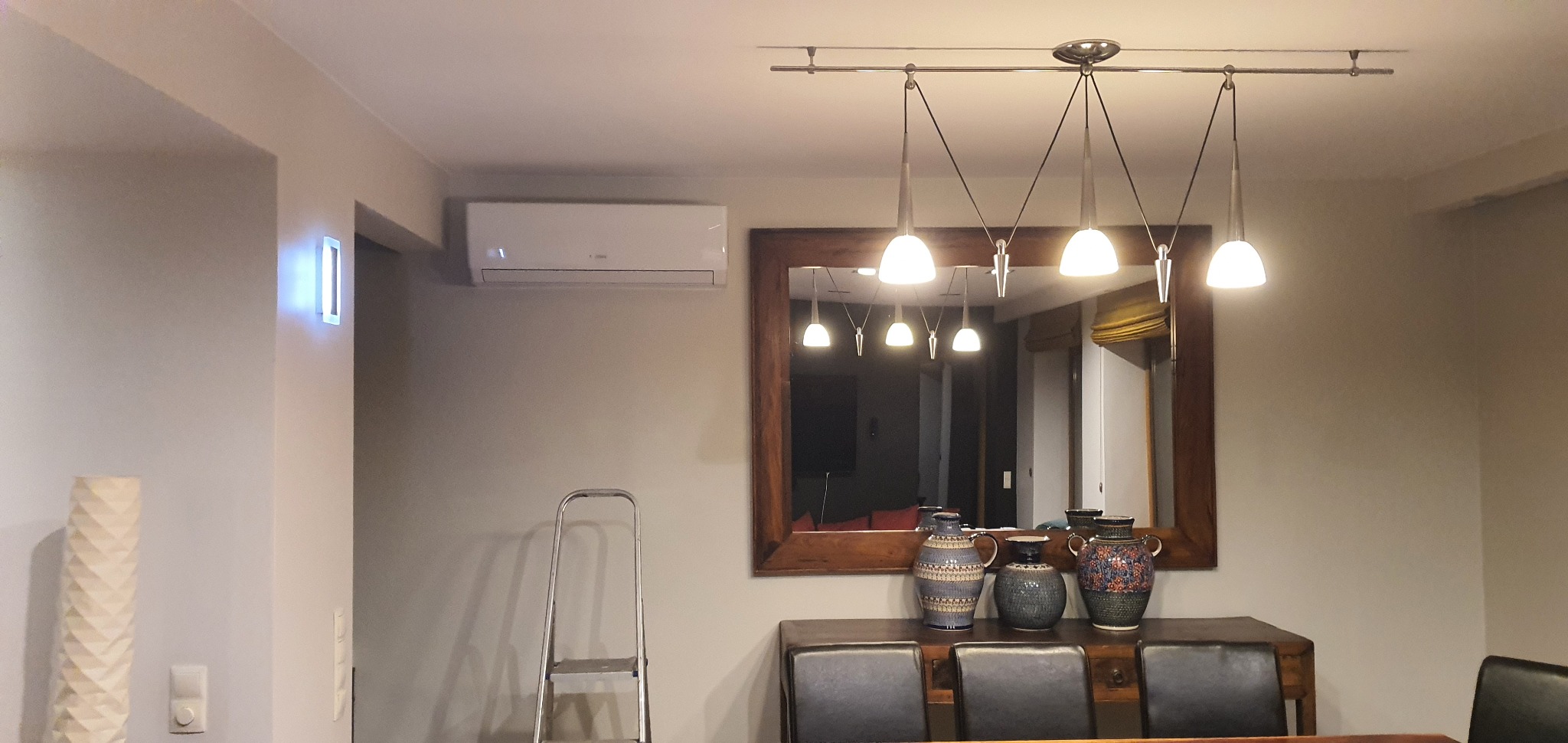 Wnętrze jadalni z zamontowaną białą klimatyzacją ścienną, widoczna drabina aluminiowa, lustro w drewnianej ramie, modernistyczna lampa sufitowa oraz dekoracyjne wazony na komodzie.
