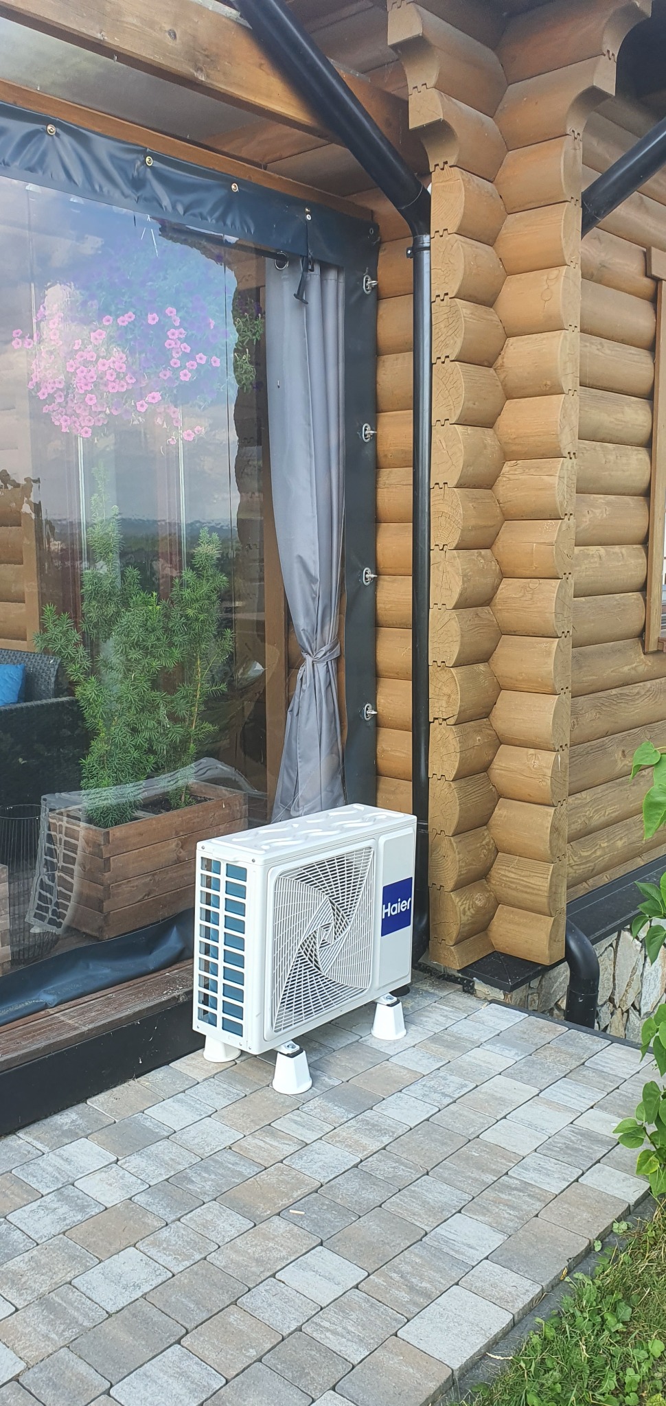 Biała jednostka zewnętrzna klimatyzatora Haier na białych podstawkach, umieszczona na kostce brukowej przy drewnianym domku z oknem, w którym widać rośliny i zasłony.