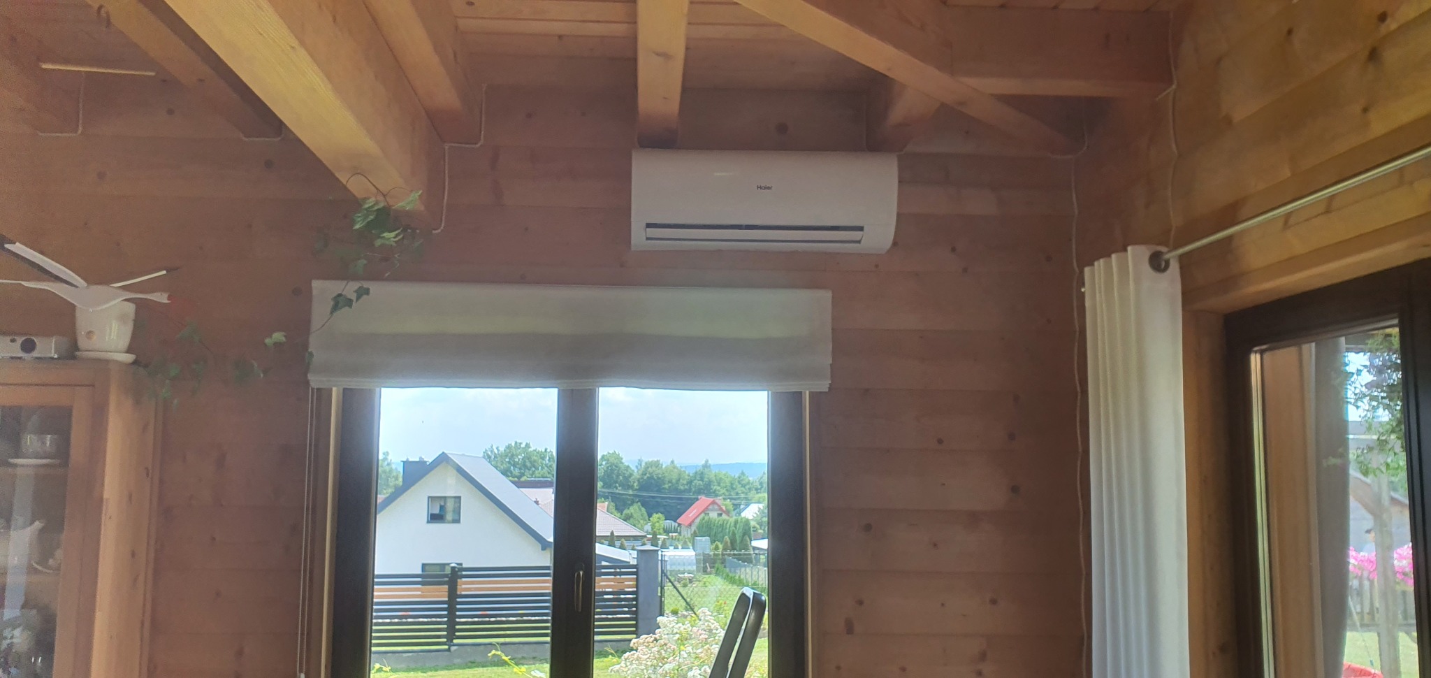 Biały klimatyzator Haier zamontowany na drewnianej ścianie w domu, widok z okna na podwórko i domy sąsiadów.