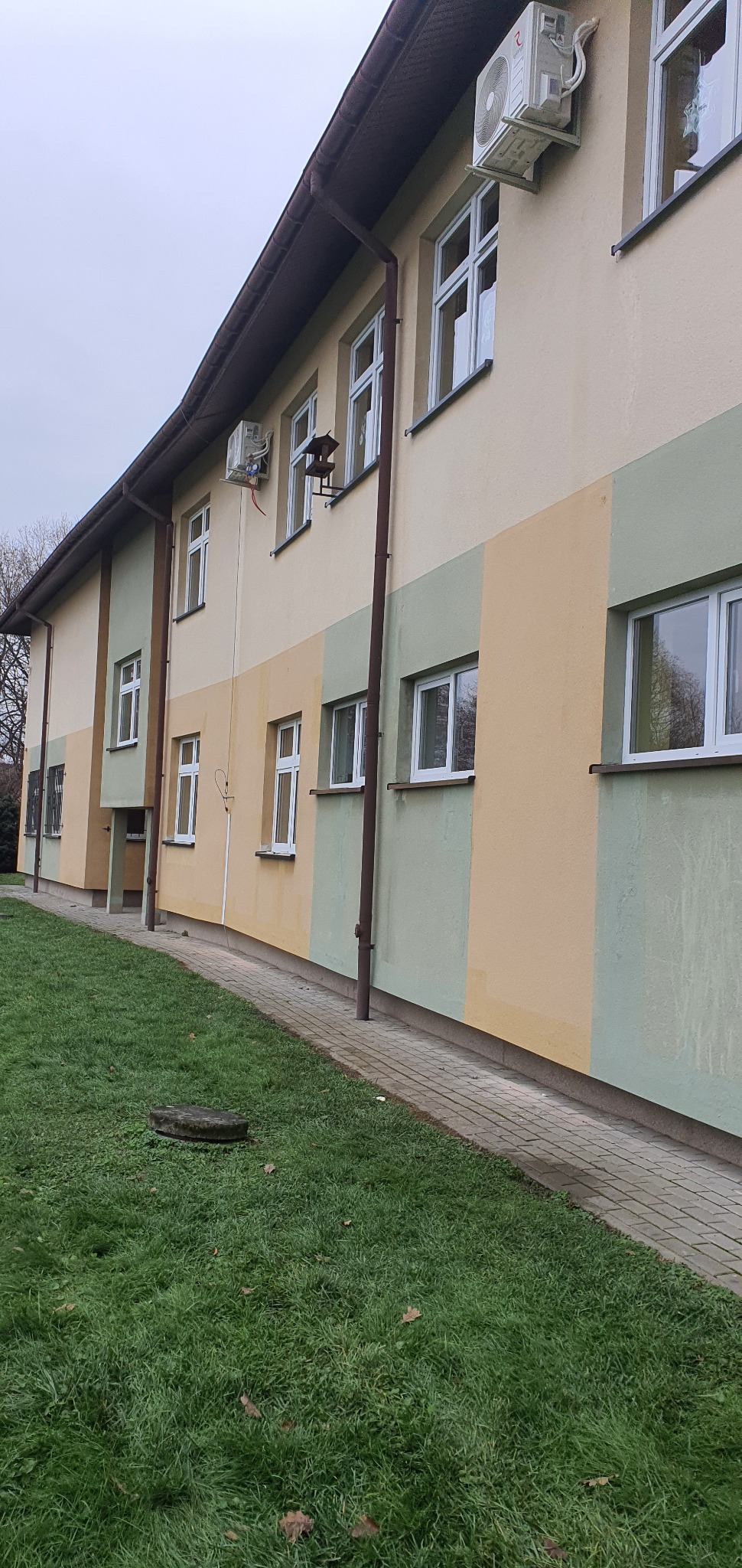 Zewnętrzne jednostki klimatyzacyjne zamontowane na elewacji budynku pomalowanej w pasy w odcieniach beżu i zieleni. Widoczne okna z białymi ramami, rynny i trawnik.