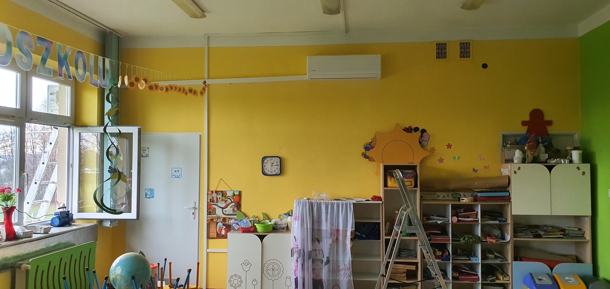 Wnętrze sali przedszkolnej z żółtymi ścianami, klimatyzatorem na ścianie, dekoracjami i meblami dziecięcymi, drabina rozstawiona w pobliżu regałów.