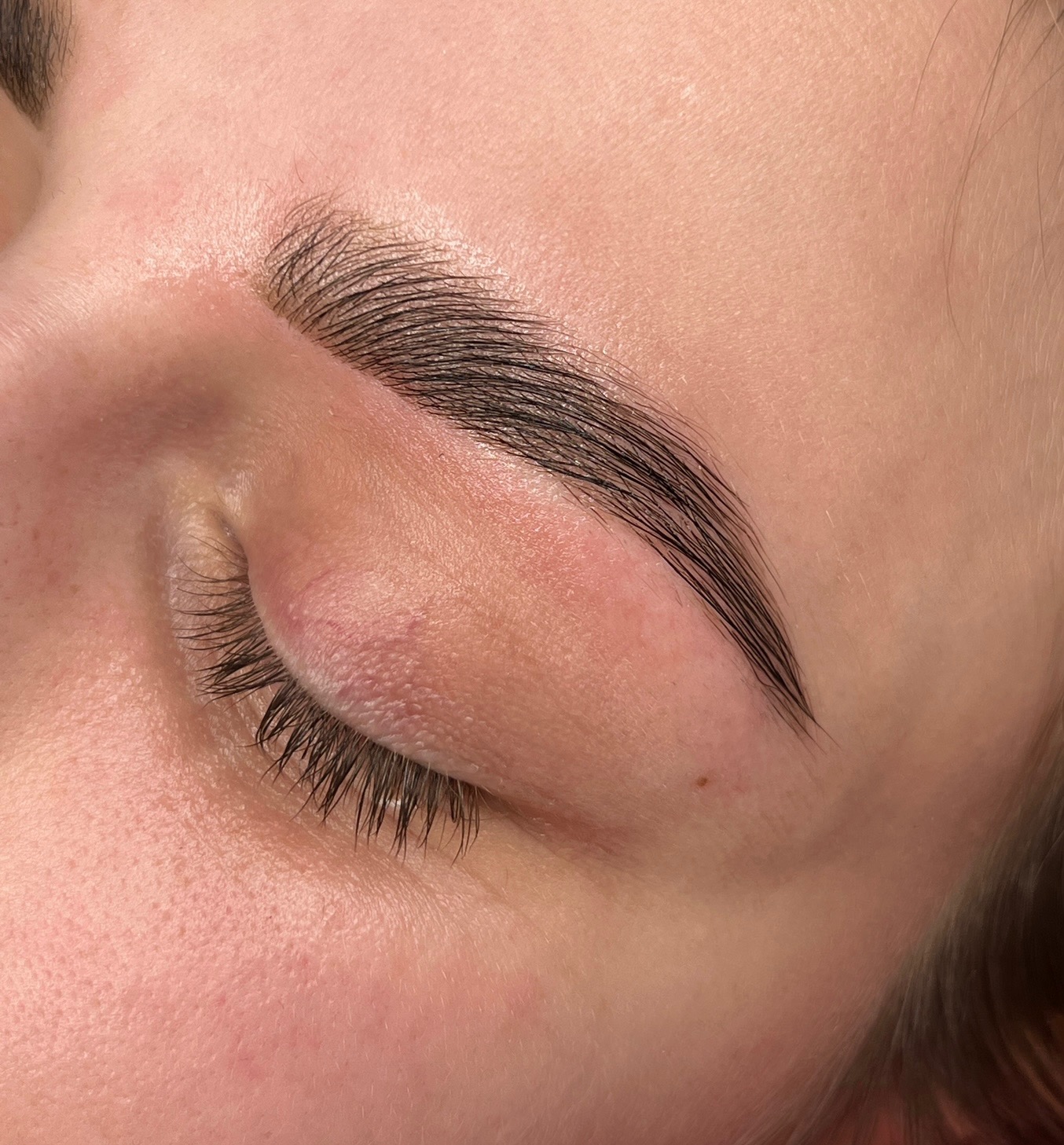 Zamknięte oko kobiety z idealnie wystylizowanymi, ciemnymi brwiami po zabiegu laminacji, widoczna gładka skóra i pojedynczy pieprzyk.