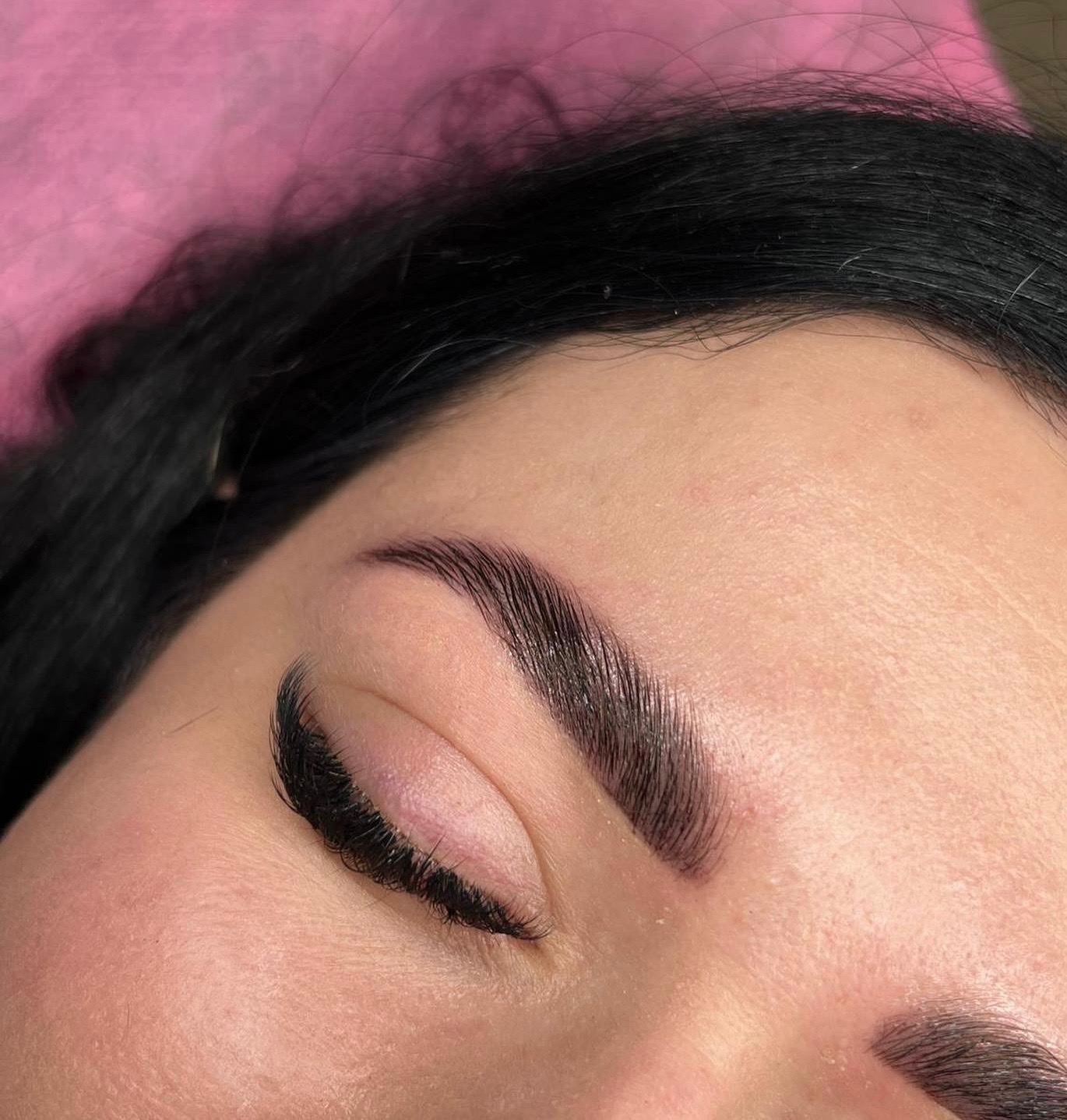 Zbliżenie na oko z ciemnymi, świeżo wystylizowanymi brwiami i przedłużonymi rzęsami na tle różowego podkładu.