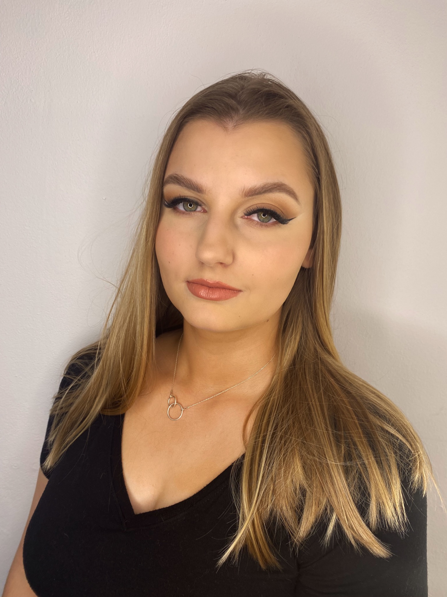 Portret młodej kobiety z profesjonalnym makijażem oczu, podkreślonym eyelinerem i delikatnymi, beżowymi ustami, na tle jasnej ściany.