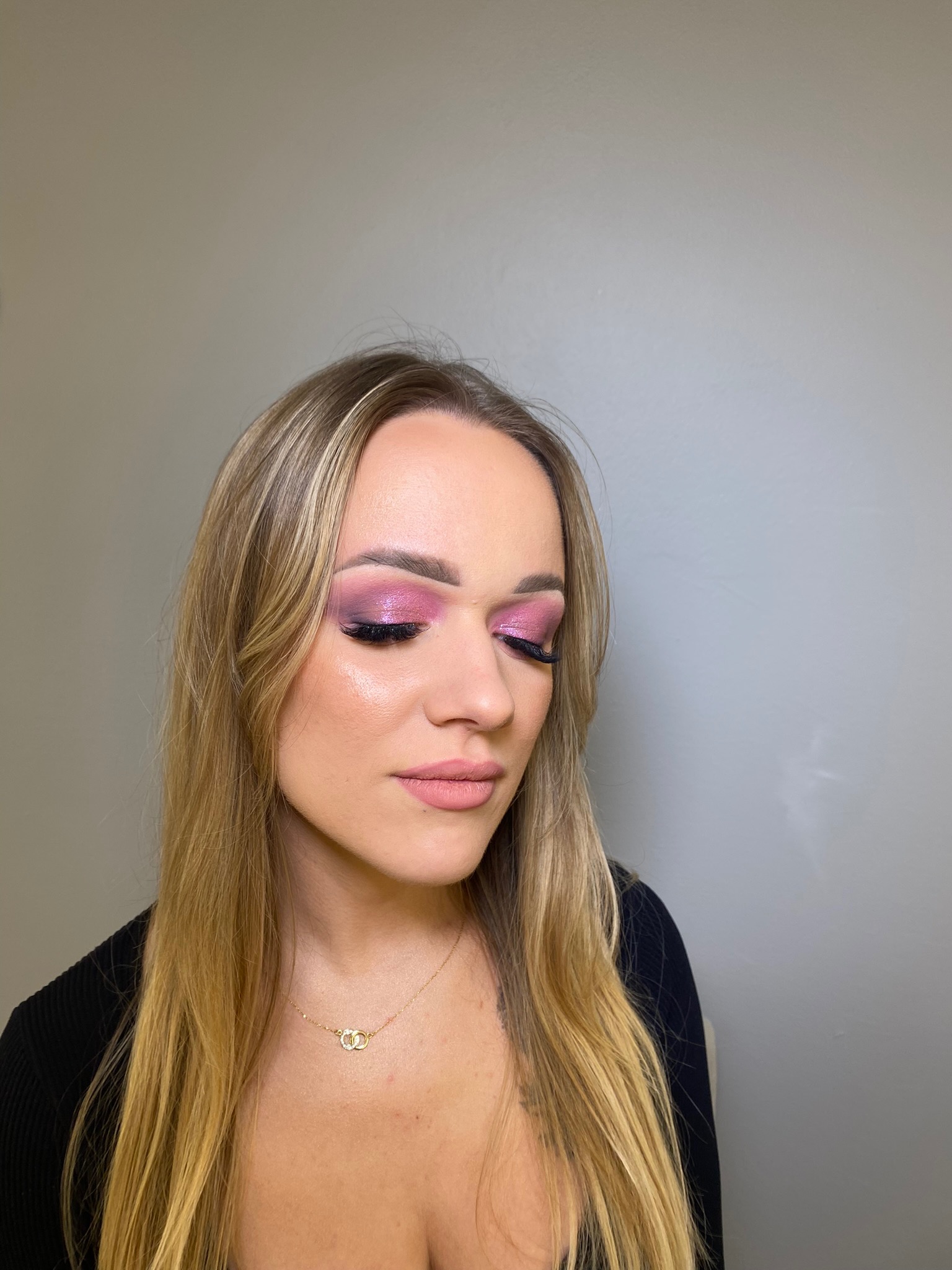 Portret kobiety z zamkniętymi oczami prezentującej różowy, błyszczący makijaż oczu i subtelny makijaż ust, z długimi blond włosami i złotym naszyjnikiem na tle szarej ściany.