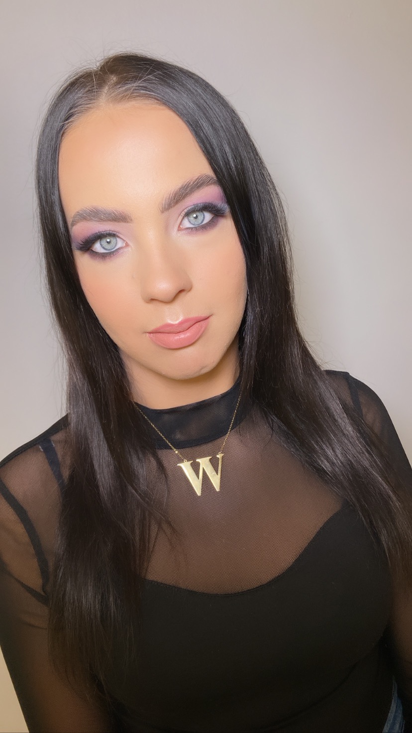 Portret kobiety z delikatnym, fioletowo-różowym makijażem oczu, błyszczącymi ustami i złotym naszyjnikiem z literą 'W' na tle jasnej ściany.