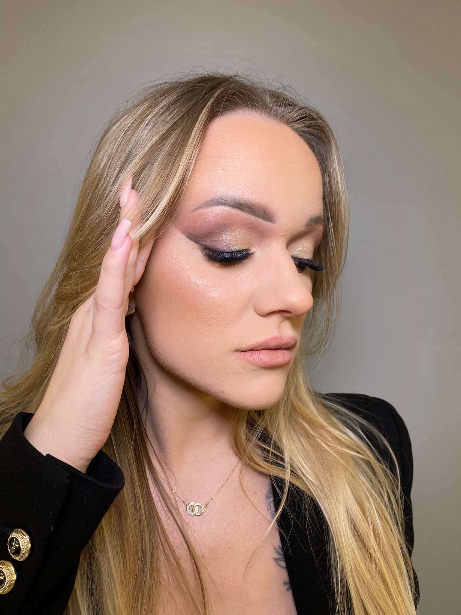 Profil kobiety z profesjonalnym makijażem, złote cienie z brokatem, czarny eyeliner i sztuczne rzęsy, blond włosy, czarny żakiet i złoty naszyjnik.