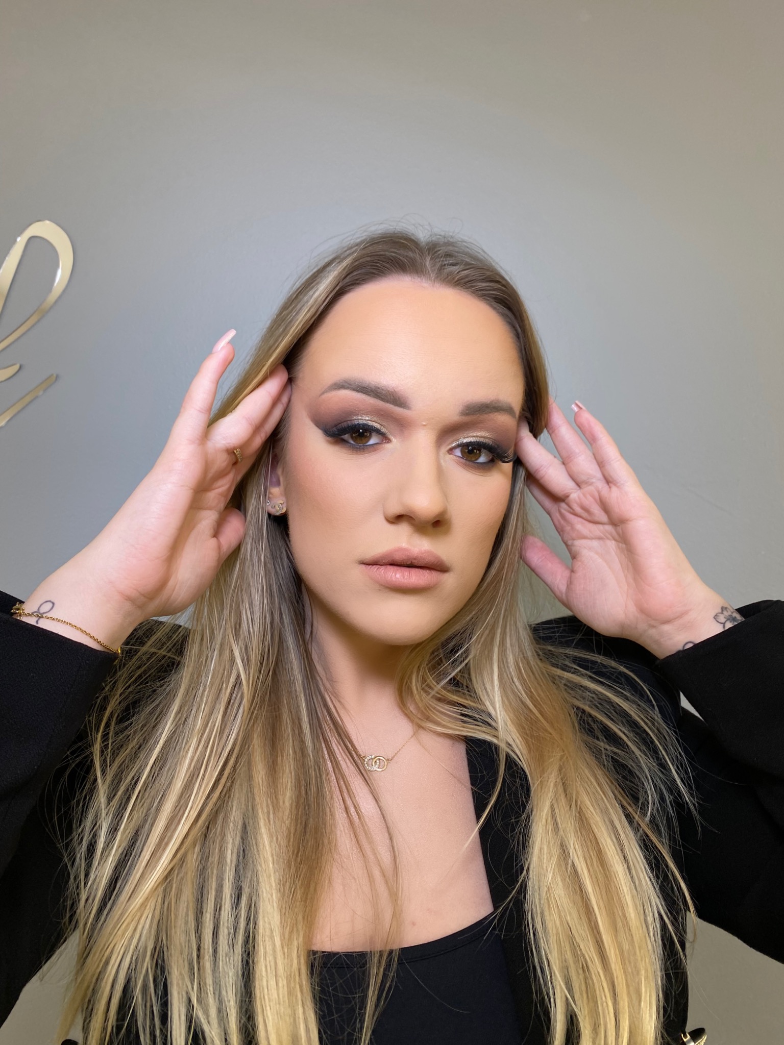 Portret kobiety z wyrazistym makijażem oczu w odcieniach brązu i złota, blond włosy opadające na ramiona, delikatna biżuteria, dłonie uniesione do skroni, szarym tło.