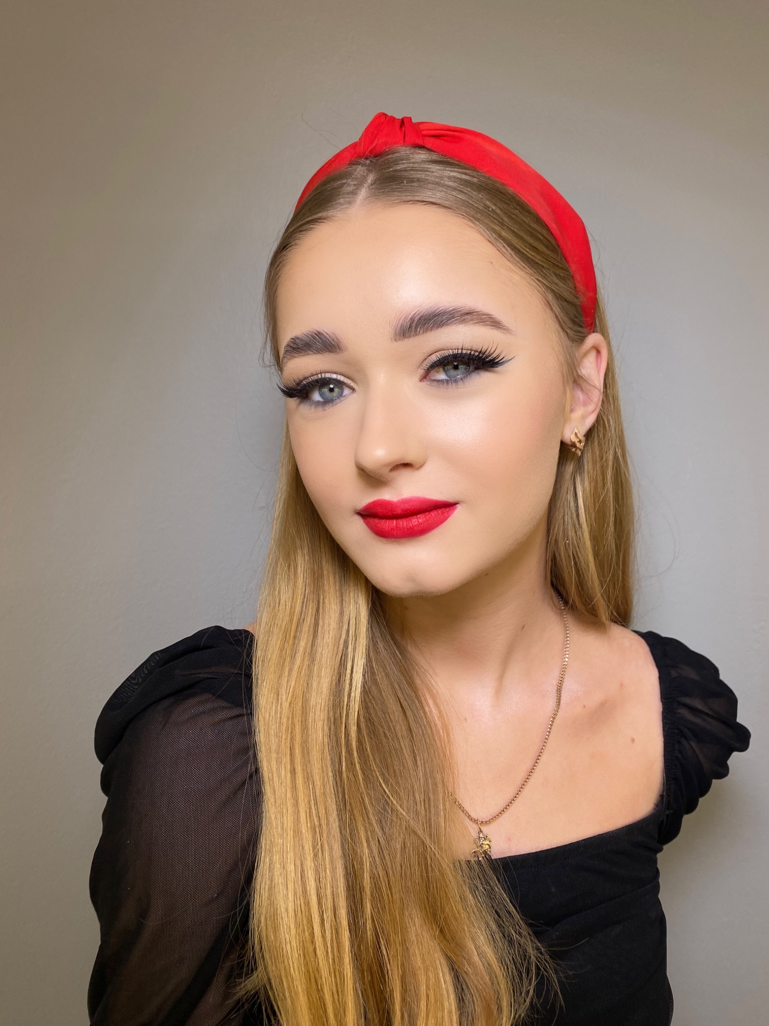 Portret młodej kobiety z blond włosami, czerwonymi ustami i czarną kreską na oku, w czerwonej opasce na głowie i czarnej bluzce z siateczki, pozującej na tle szarej ściany.