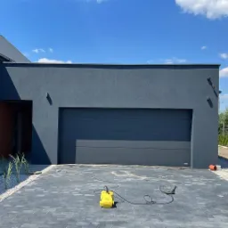 Nowoczesny dom z szarą elewacją i bramą garażową, widoczny fragment kostki brukowej i myjka ciśnieniowa, w tle błękitne niebo z chmurami.