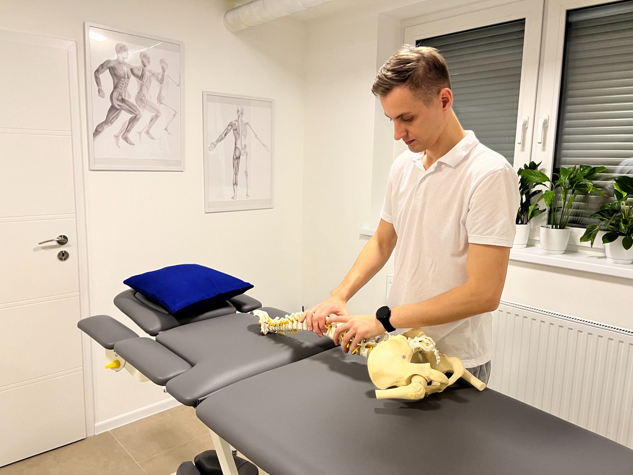 Fizjoterapeuta demonstruje budowę kręgosłupa i miednicy na modelu anatomicznym, leżącym na szarym stole rehabilitacyjnym w jasnym gabinecie, z plakatami przedstawiającymi ludzki szkielet i mięśnie...