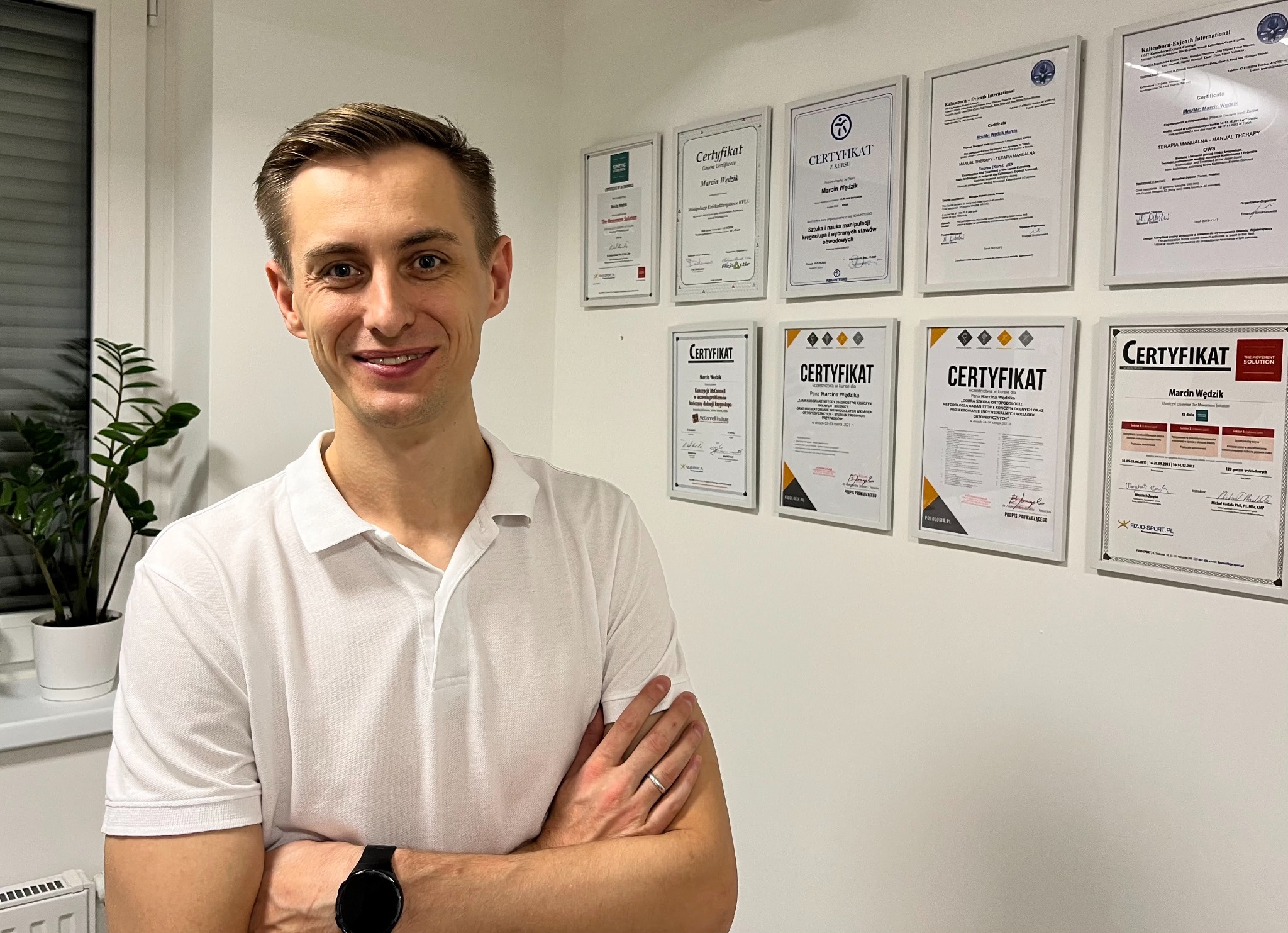 Uśmiechnięty mężczyzna w białej koszulce polo, z zegarkiem na nadgarstku i obrączką, stojący na tle ściany z powieszonymi certyfikatami ukończenia kursów i szkoleń oraz rośliną doniczkową przy oknie.