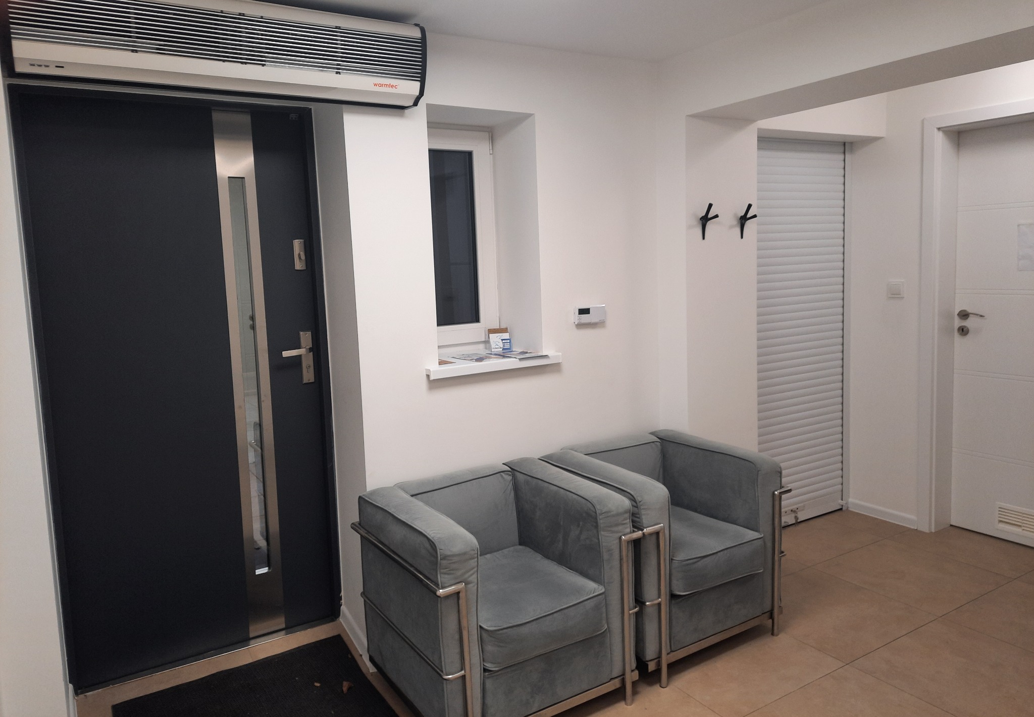 Poczekalnia z dwoma szarymi fotelami na metalowych nogach, czarnymi wieszakami na ścianie i oknem z białym parapetem.