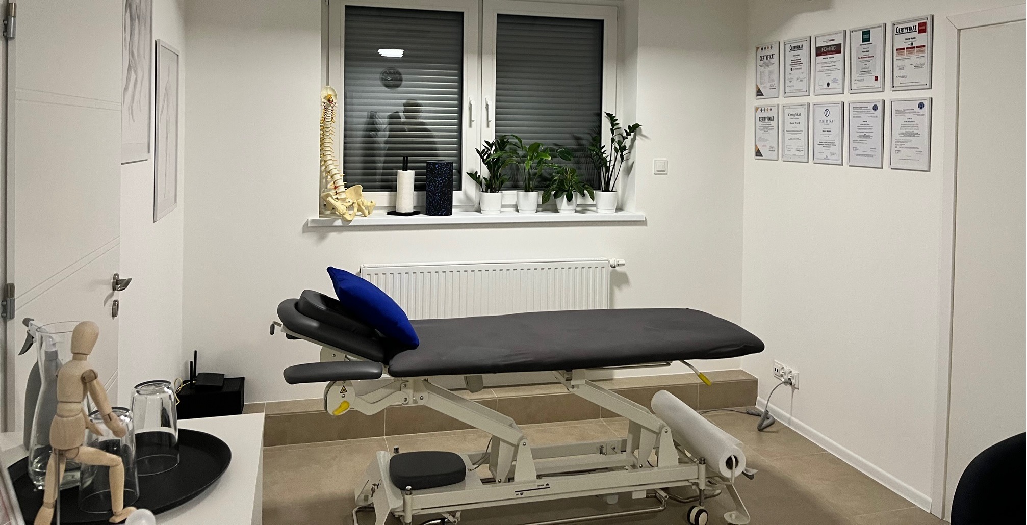 Jasne wnętrze gabinetu z profesjonalnym stołem do zabiegów, modelem kręgosłupa na parapecie, licznymi certyfikatami na ścianie i drewnianą figurką człowieka na stoliku.