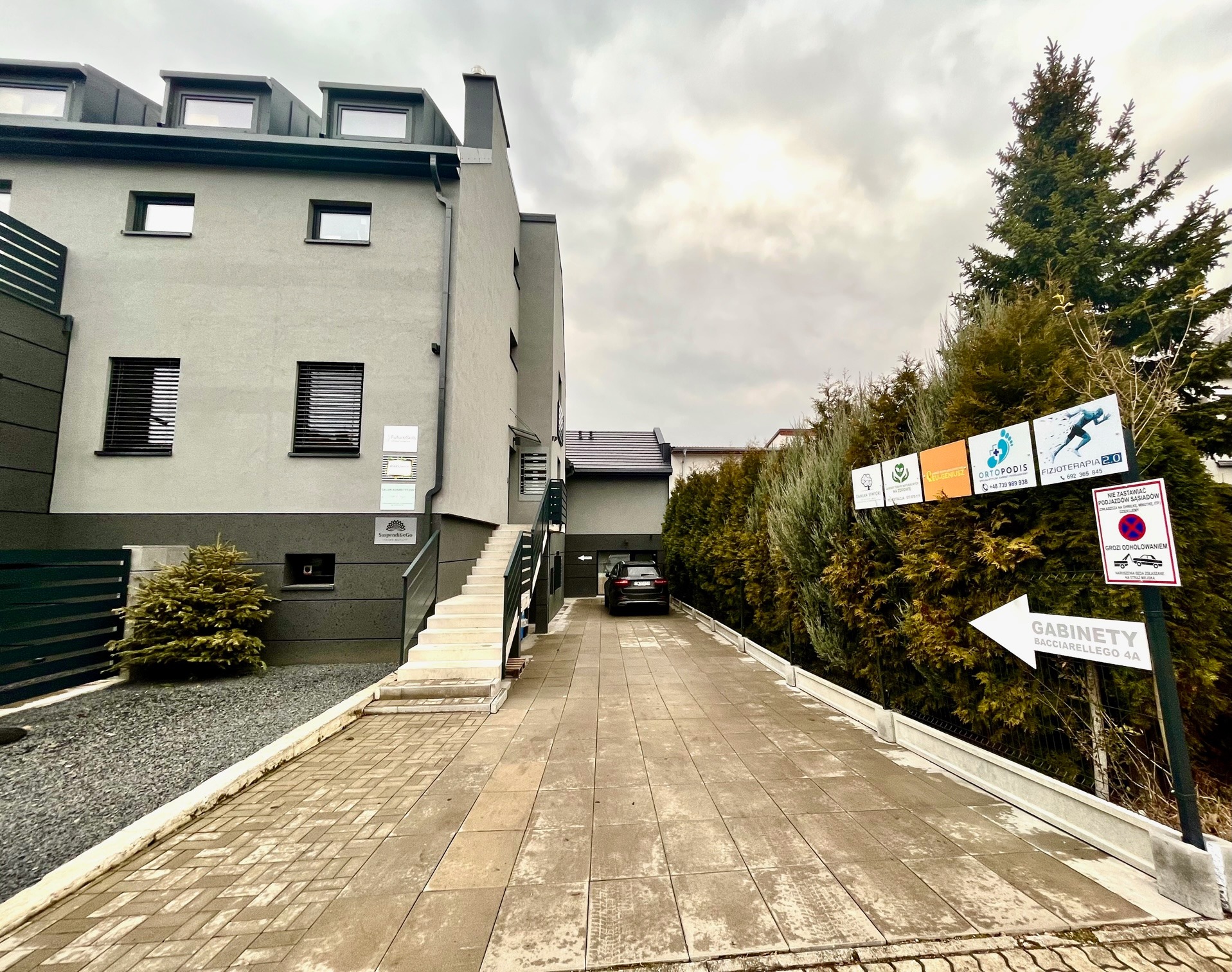 Elewacja budynku z gabinetami, tabliczki informacyjne na elewacji, dojście wyłożone kostką, znak z napisem 'Gabinety Bacciarellego 4A' oraz tablice reklamowe z informacjami o usługach, w tle...