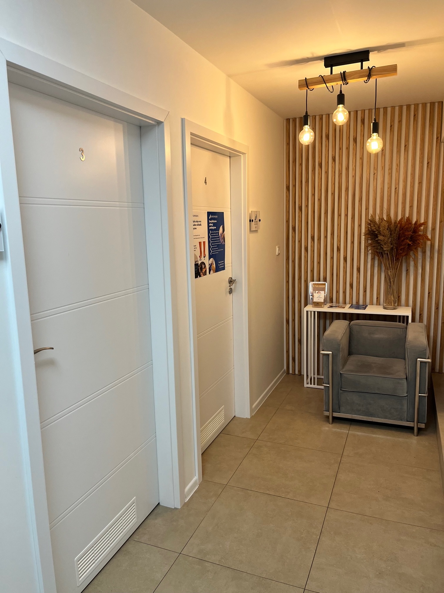 Poczekalnia w gabinecie: Dwoje białych drzwi z numerami 3 i 4, szary fotel, drewniana ściana z dekoracyjnymi suszonymi kwiatami i lampami w stylu retro.