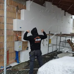 Pracownik w bluzie z logo firmy ES-BUD prezentuje fragment ocieplonej elewacji styropianem w trakcie opadów śniegu, widoczna skrzynka elektryczna i rusztowanie.