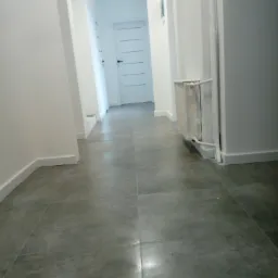 Korytarz z szarymi płytkami podłogowymi, białymi ścianami i grzejnikiem. W tle białe drzwi z poziomymi wgłębieniami.