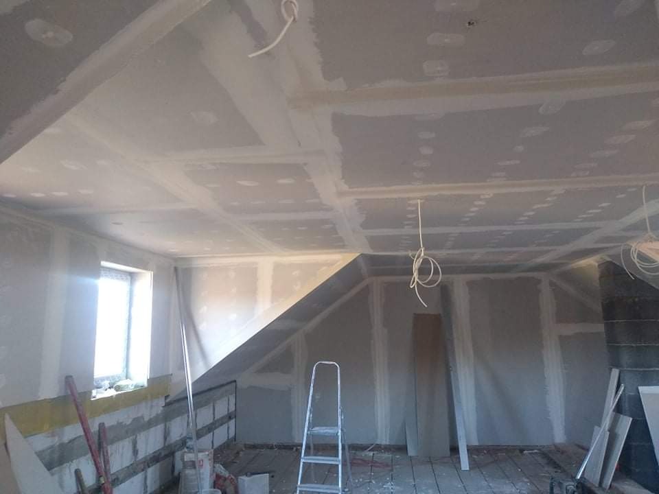 Poddasze w trakcie wykańczania płytami gipsowo-kartonowymi, widoczna drabina, okno i częściowo ocieplone ściany, przewody elektryczne zwisające z sufitu.