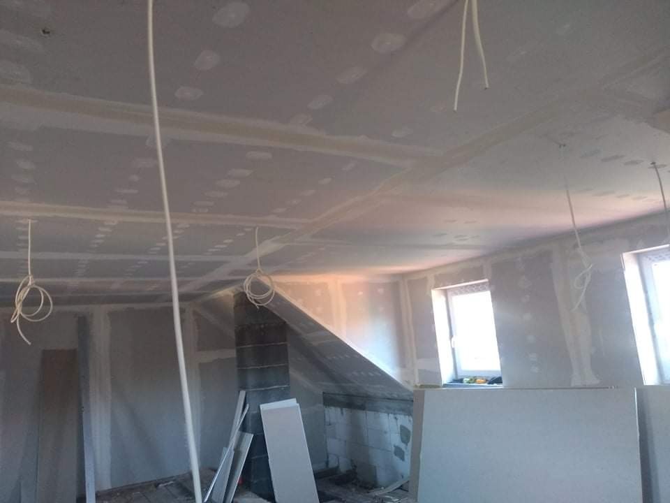 Wnętrze w trakcie montażu płyt gipsowo-kartonowych na ścianach i suficie, widoczne zwisające kable elektryczne, surowy komin i okna w tle.